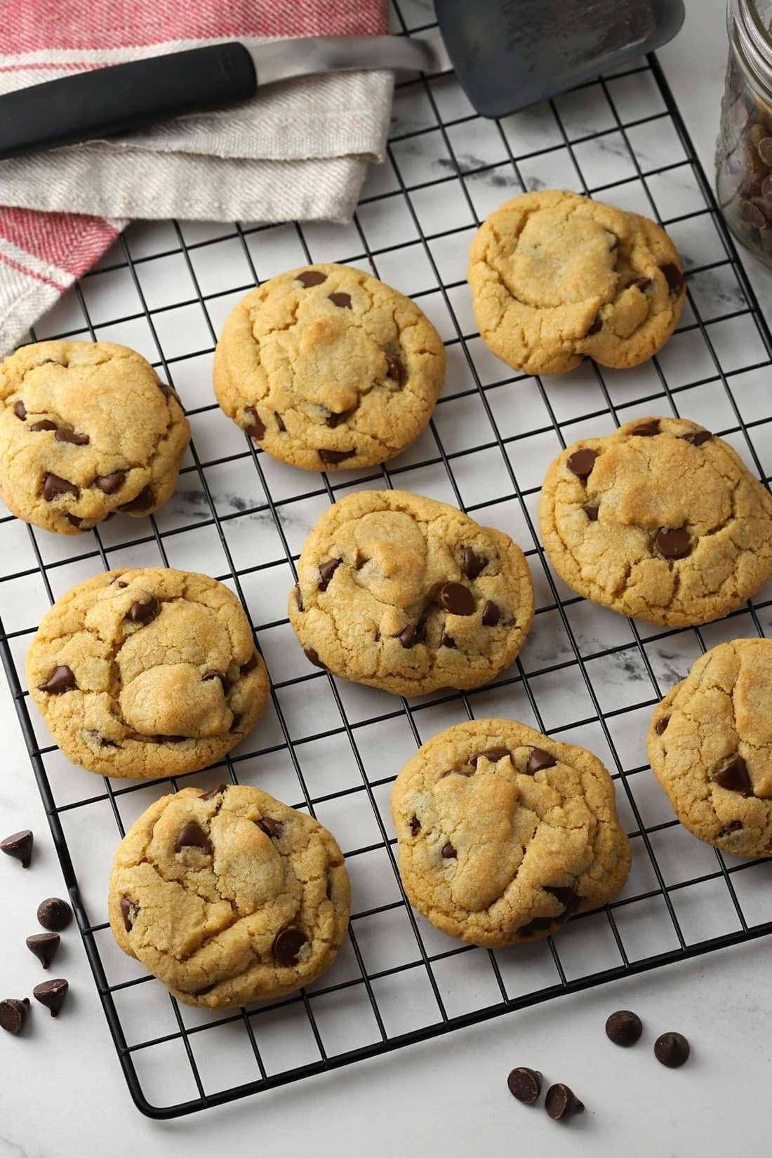 Fresh golden chocolate chip cookies cooling on a black wire rack, marble surface, striped towel and spatula ne