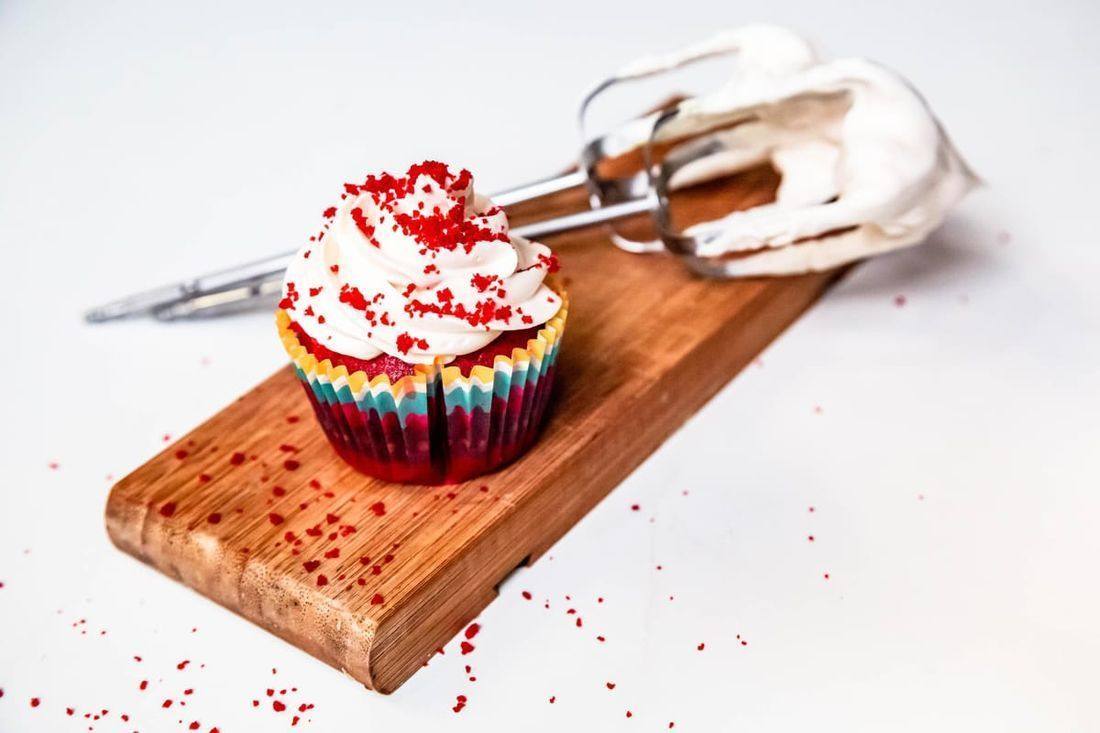 A red velvet cupcake with white frosting and red crumbs sits on a wooden board beside a frosting-covered mixer