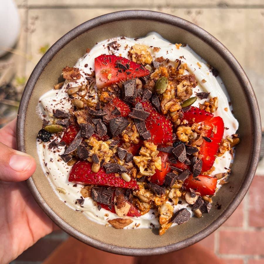 A hand holds a bowl of yogurt topped with fresh strawberries, granola, seeds, and chocolate shavings; high cla
