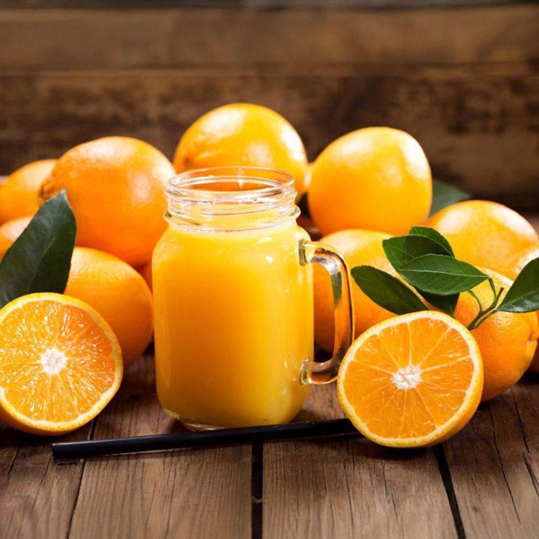 A mason jar mug of fresh orange juice surrounded by whole and halved oranges, leaves, on rustic wooden table.