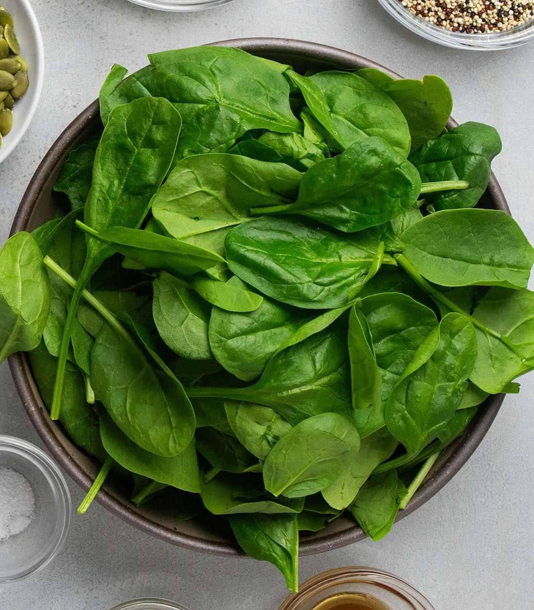 A brown bowl filled with fresh green spinach leaves, surrounded by small bowls of seeds, quinoa, and salt; no